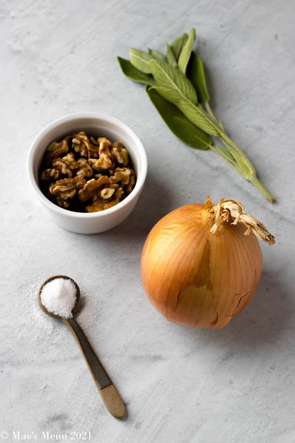 An up-close shot of an onion, cup of walnuts, sprig of sage, and small spoon of salt.