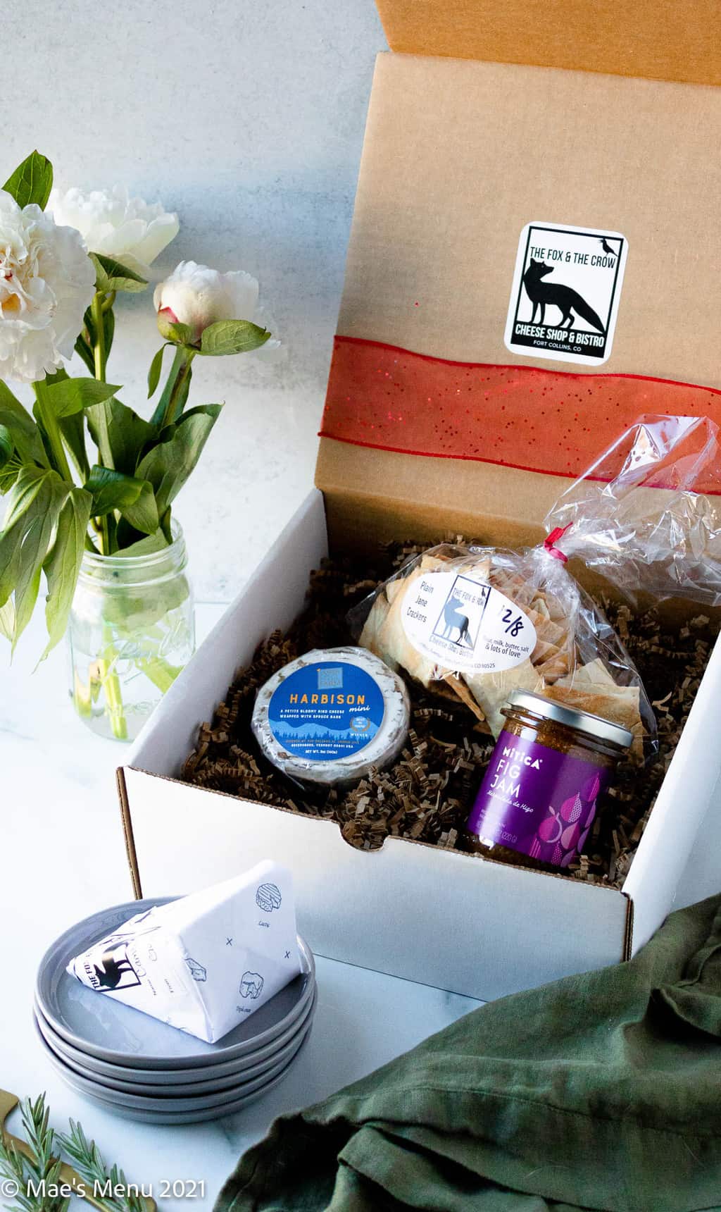A white packing box of cheese, crackers, and fix jam next to a small vase of flowers.