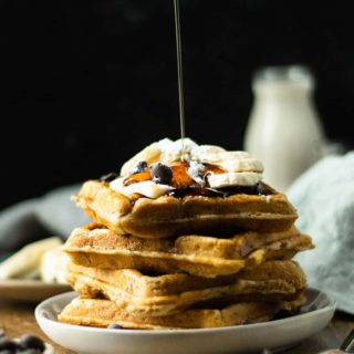 A side shot of a stack of banana waffles with syrup drizzling on them.