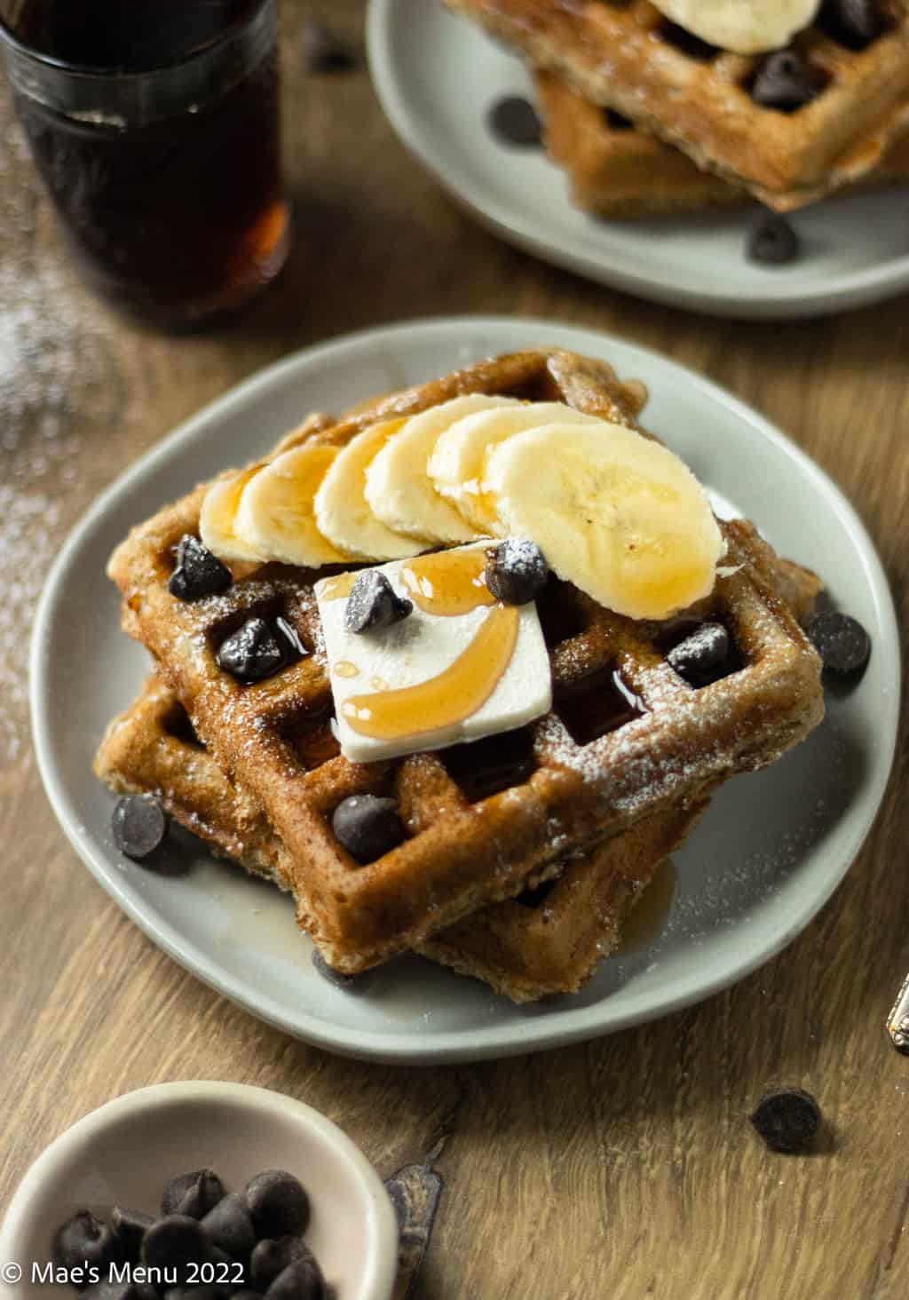 An up-close side shot of a white plate with banana waffles, bananas, syrup, and chocolate chips.