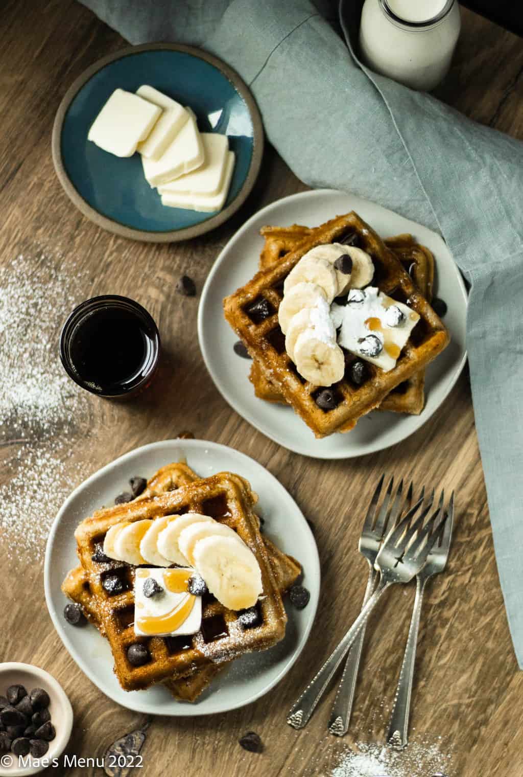 Plates of banana waffles with bananas, chocolate chips, syrup, and a small cup of syrup and milk.