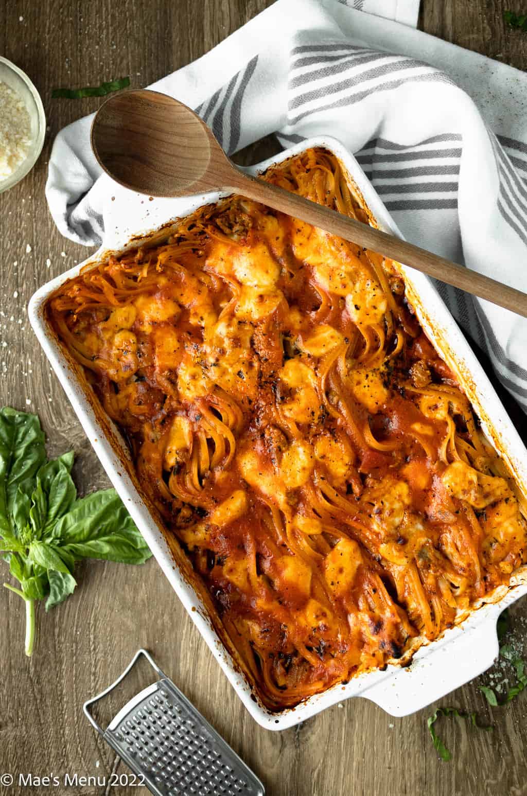 An overhead shot of a large pan of no boil pasta bake surrounded by herbs, cheese, and a wooden spoon.