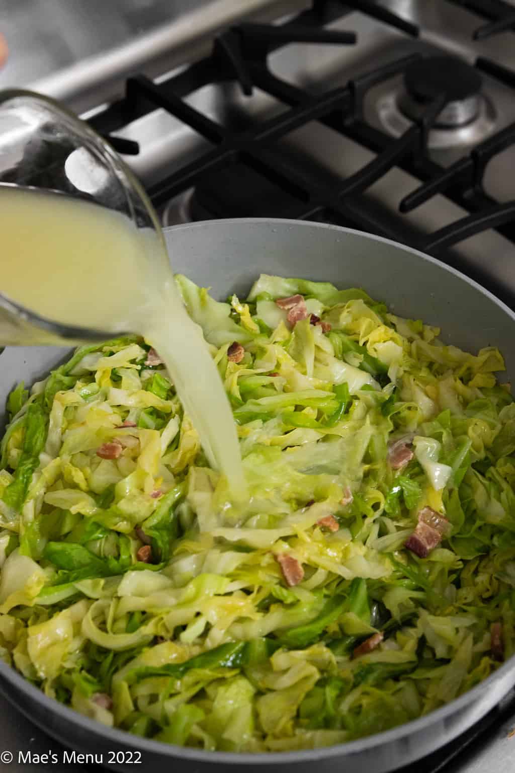 A large saute pan with chicken broth pouring into a saute pan with cabbage and bacon.