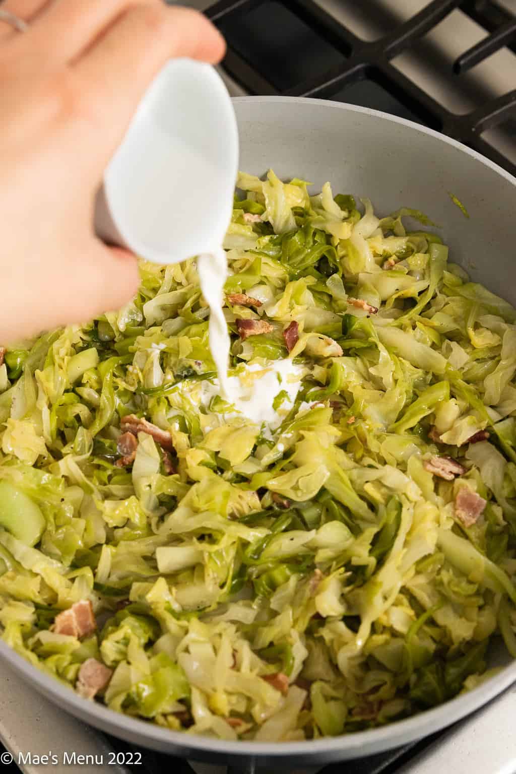 Pouring cream into a saute pan with cabbage and bacon.