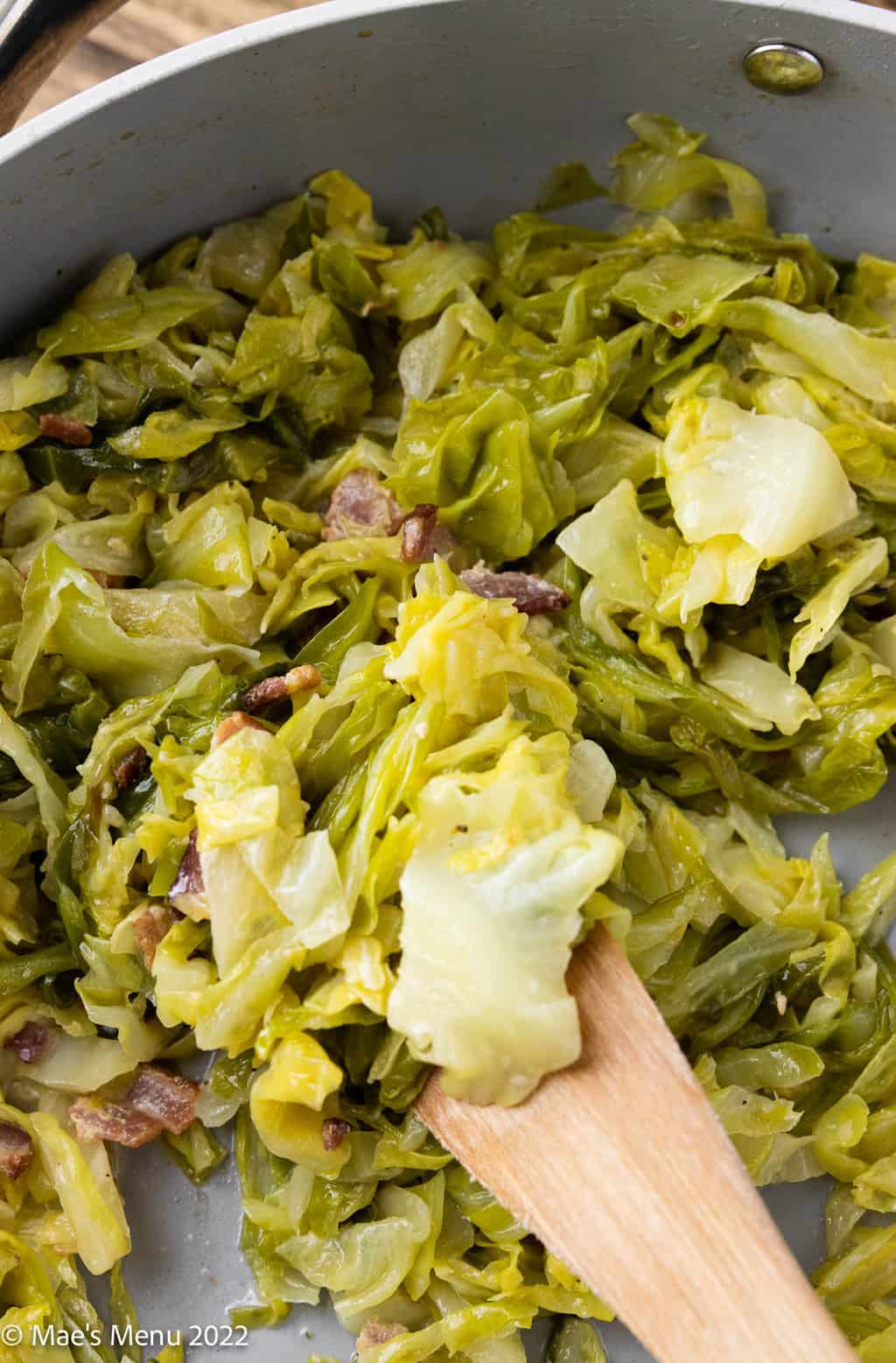 An up-close overhead shot of sautéed cabbage and bacon in a pan with a women spoon.
