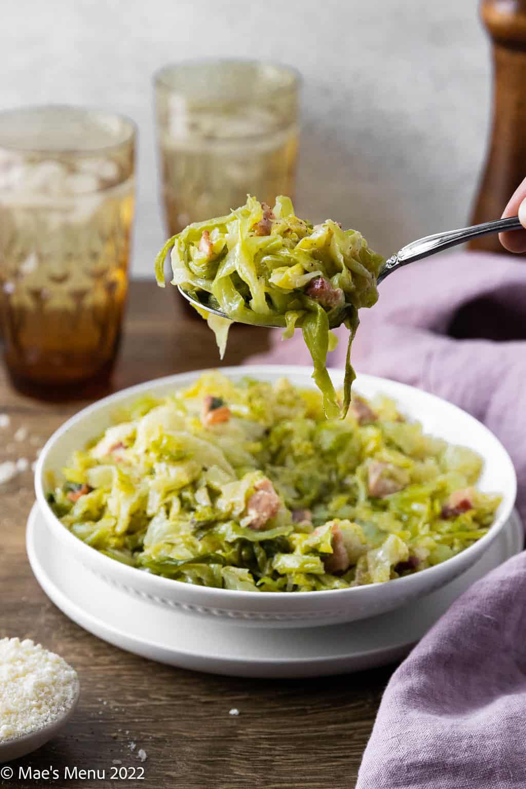 A spoonful of creamed cabbage with bacon with a bowl, glasses of water, and a pepper mill in the background.