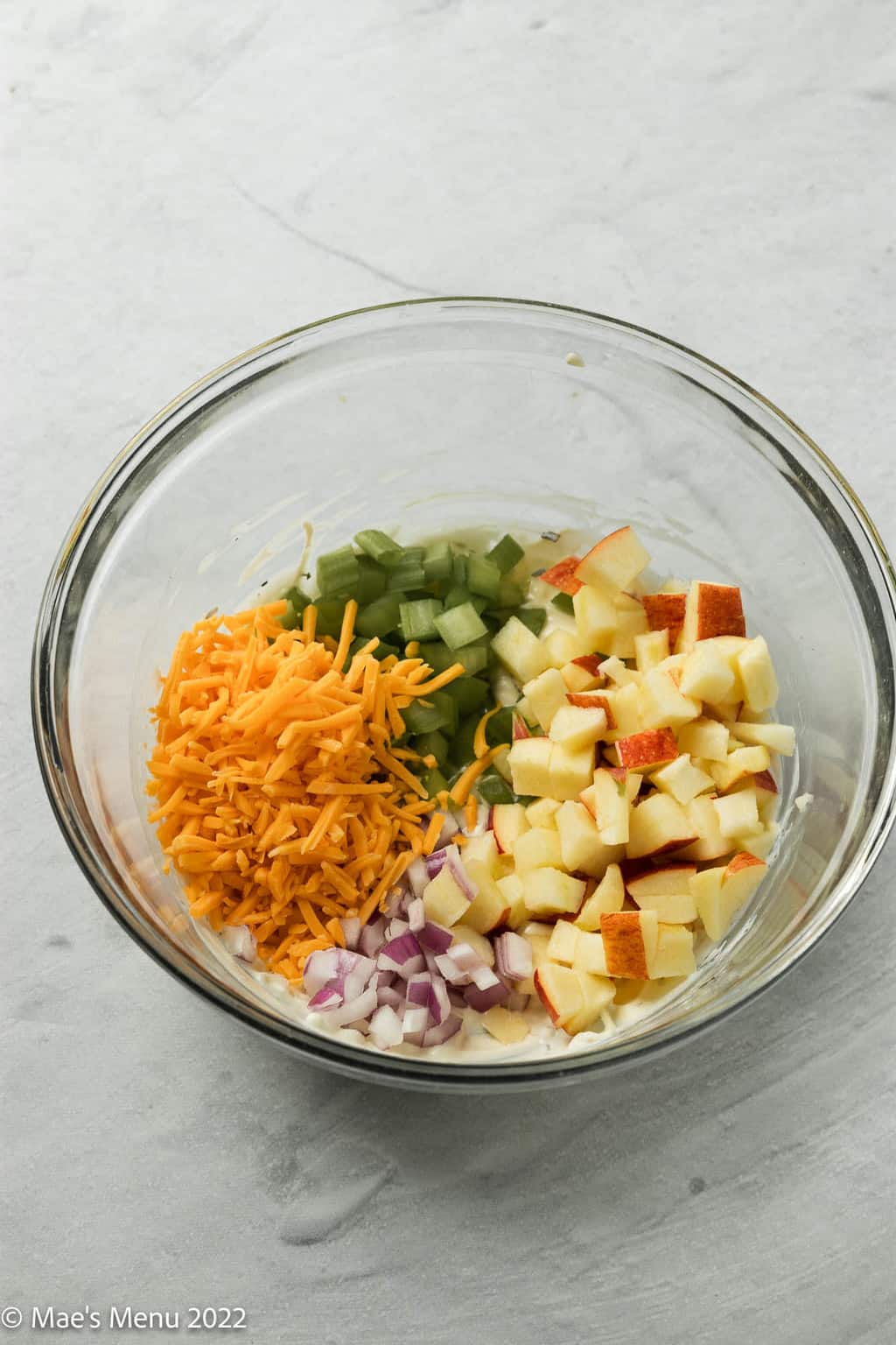 An overhead shot of a glass mixing bowl with celery, red onions, and apples, ceddar cheese.