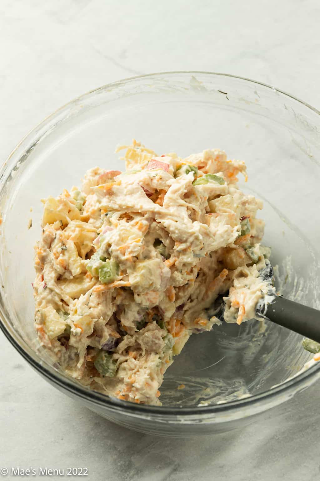 An angled shot of a large glass mixing bowl with chicken salad and a silicone spatula in it.