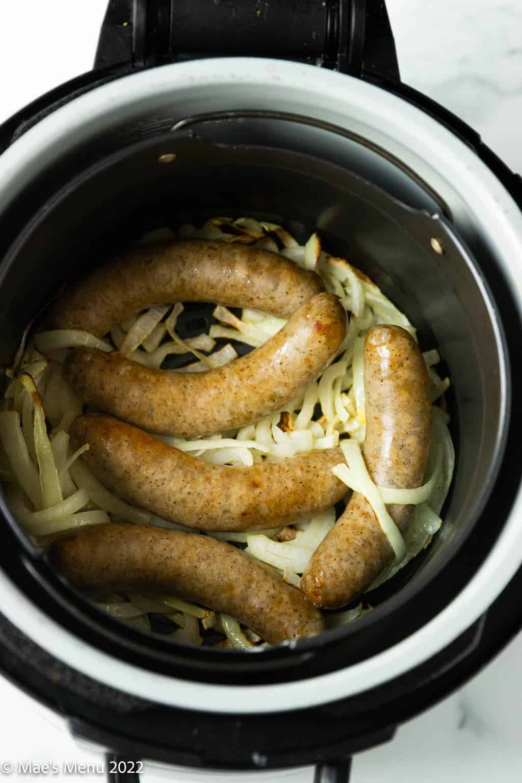 An overhead shot of onions and air fried brats in an air fryer basket.