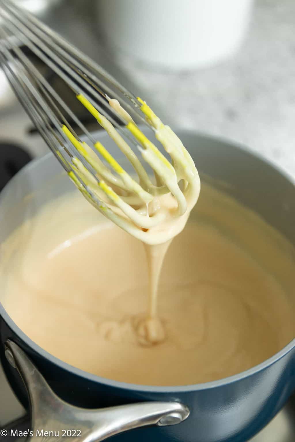 A whisk drizzling cheese sauce into a saucepan.