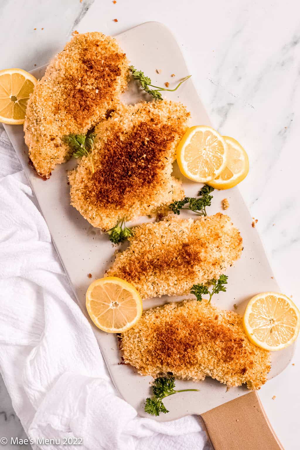 A cutting board with breaded panko chicken, lemon, and parsley on it.
