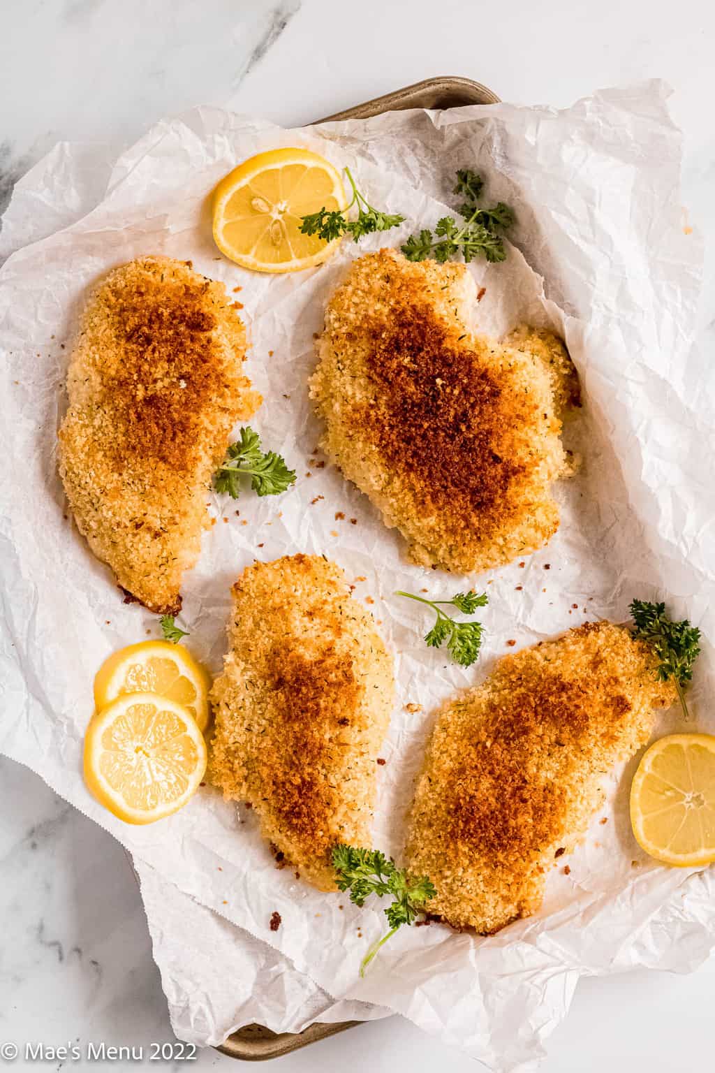 Herbed panko chicken breasts on a baking sheet with parsley and lemon.