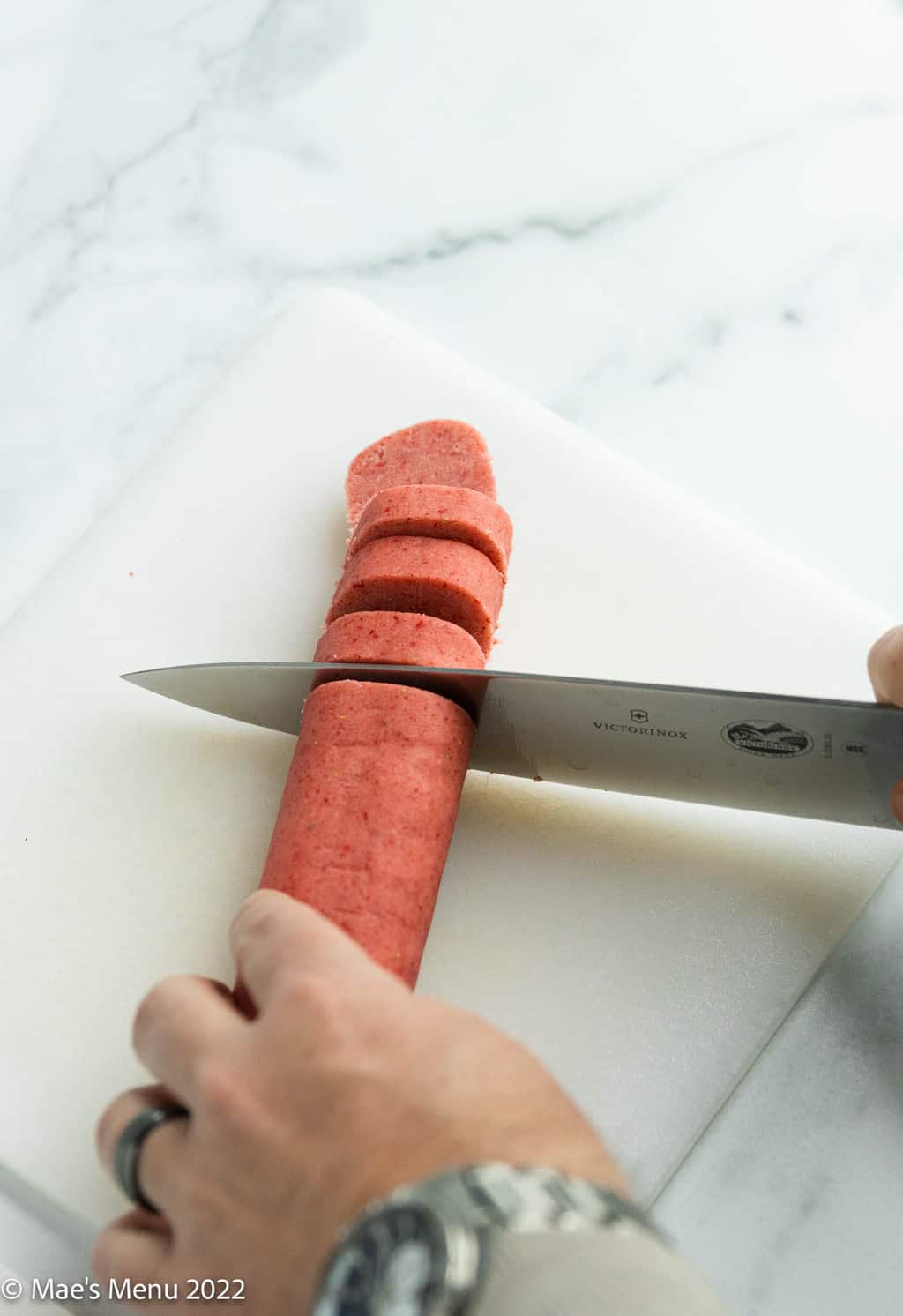 A hand slicing a cylinder of strawberry shortbread dough.