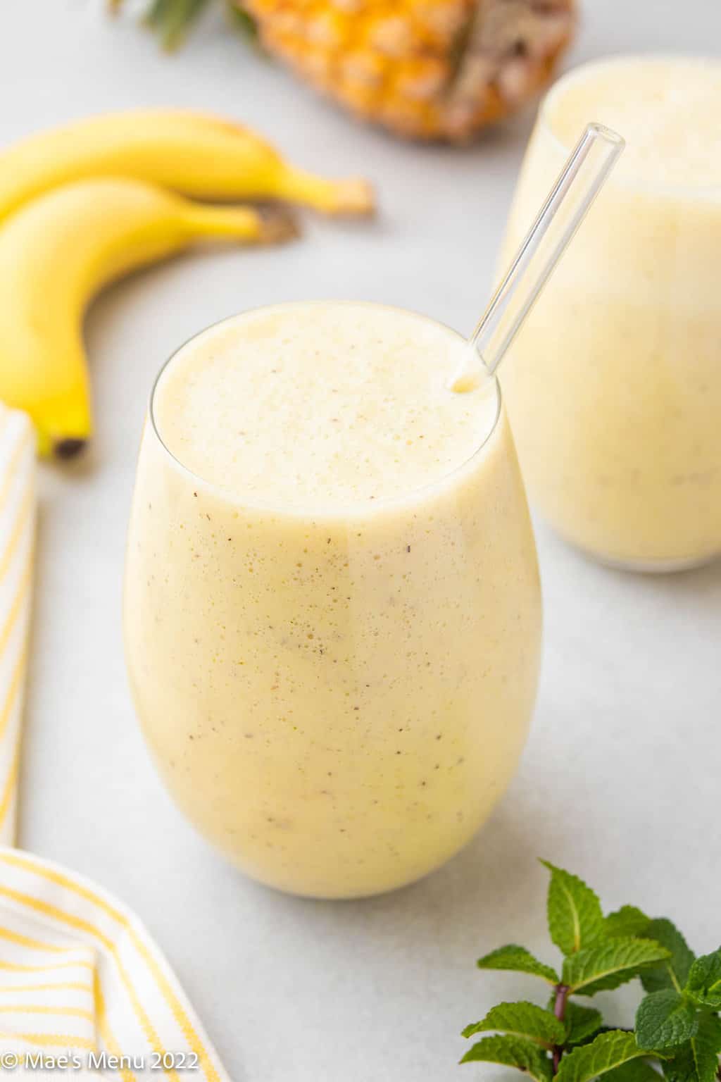 Two glasses of pineapple banana smoothie with bananas and a pineapple in the background.