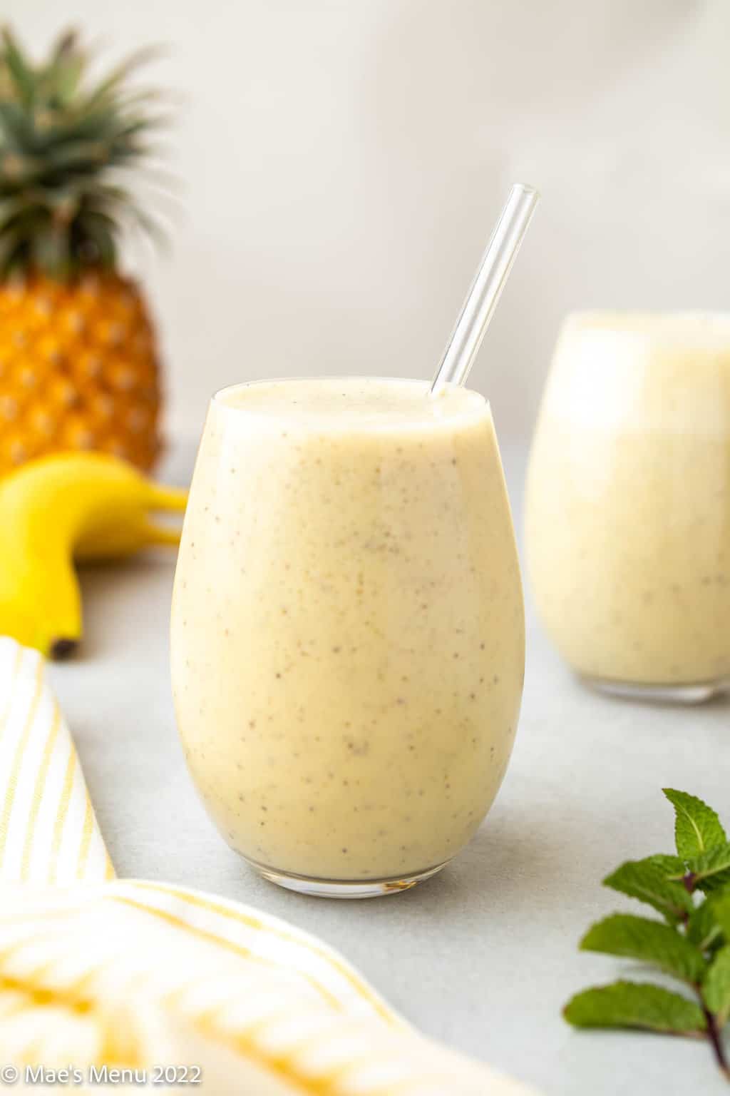 A side shot of two glasses of pineapple banana smoothie on a counter with pineapple and banana in the background.