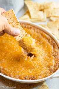A hand dipping a chip into a dish of cheesy refried bean dip.