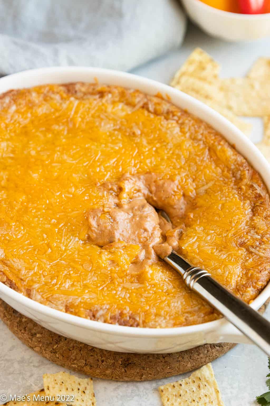 A white dish of cheesy refried bean dip with a spoon in it.