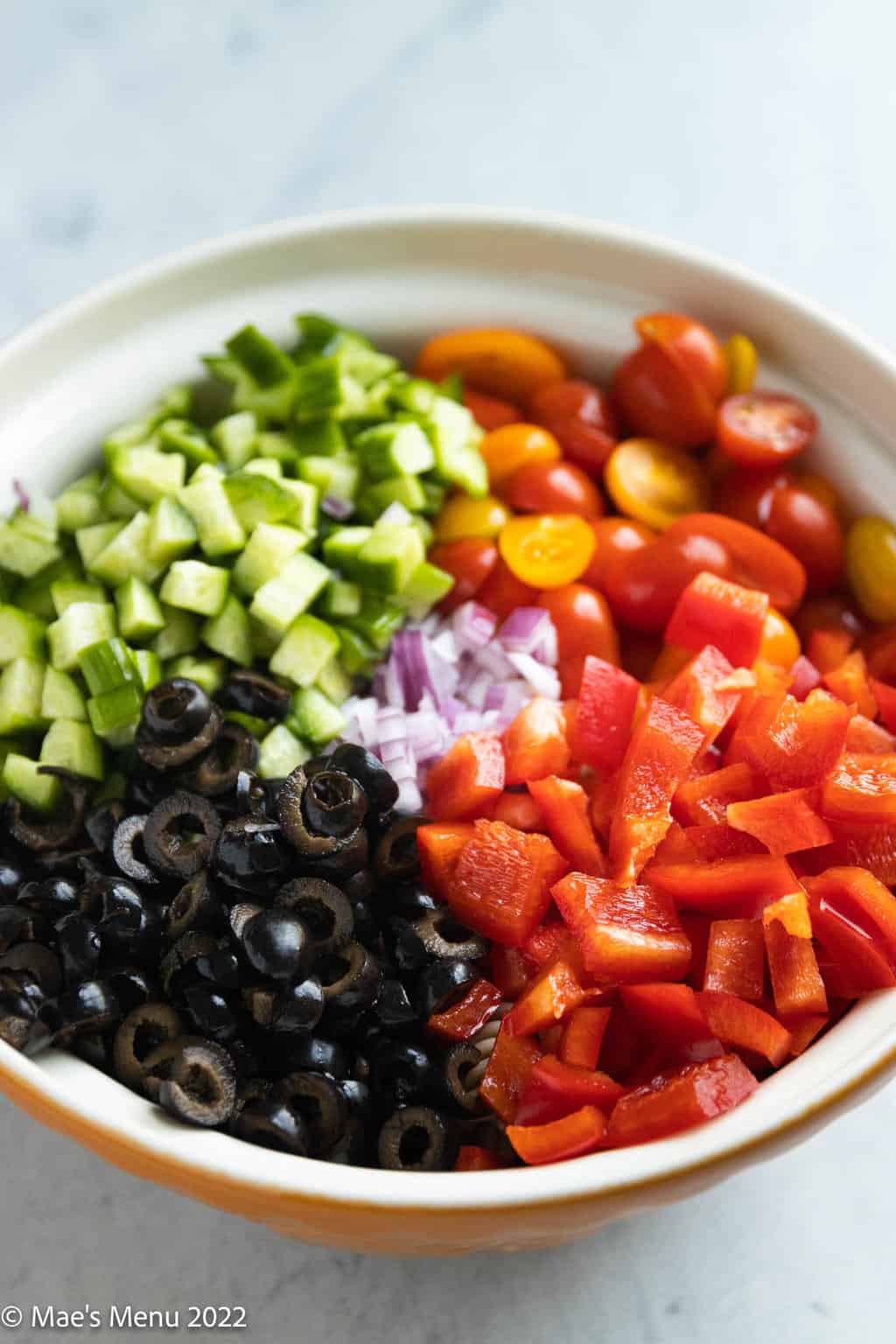 A mixing bowl with diced red peppers, red onion, black olives, cucumber, and halved cherry tomatoes.