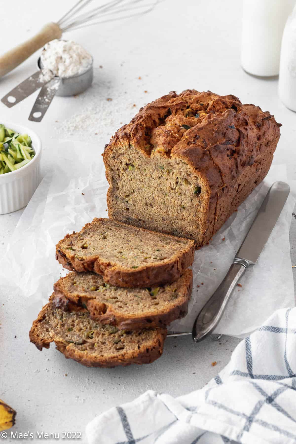 A partially sliced loaf of banana zucchini bread on a cooling rack with a knife.