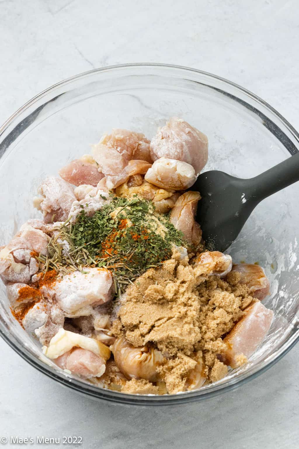 A clear mixing bowl of chicken with brown sugar, salt, dried herbs, and other seasonings.