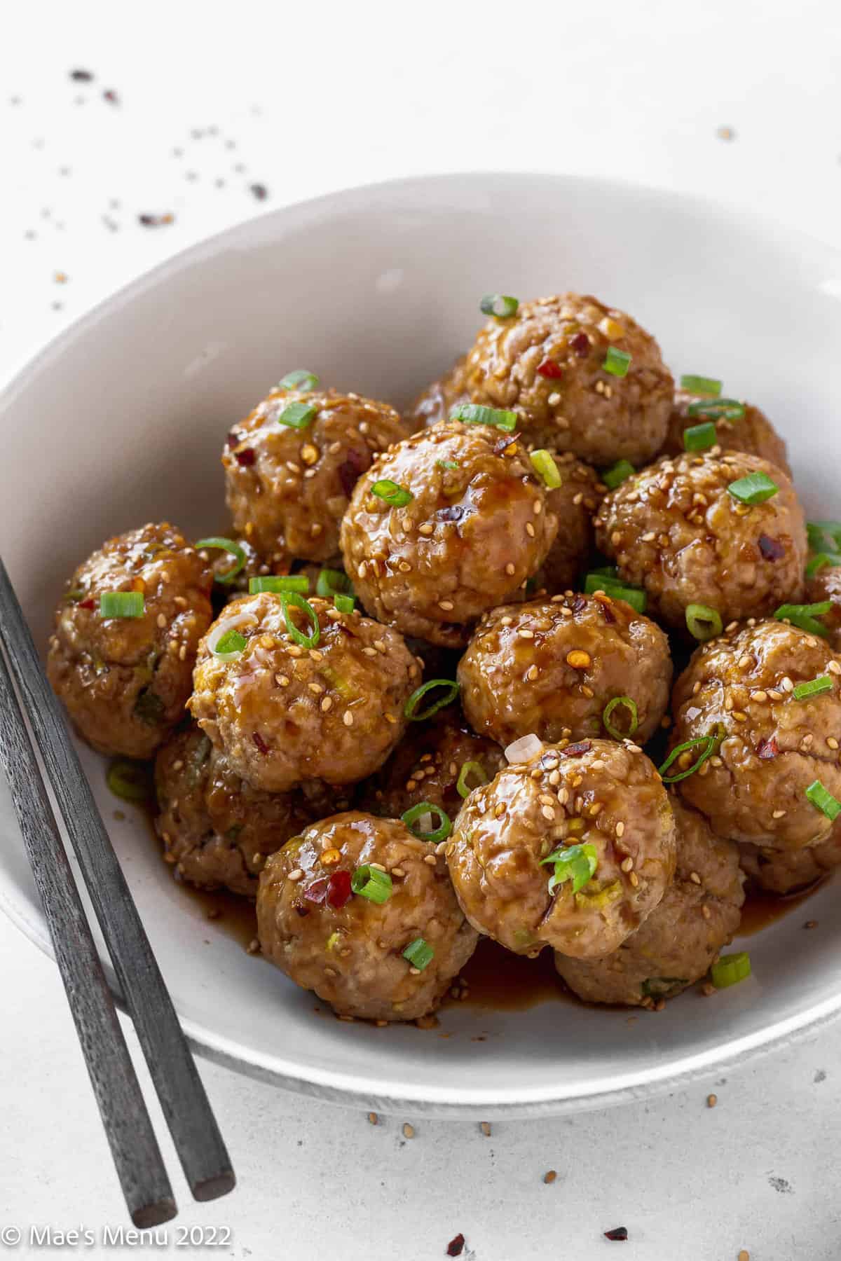 white serving bowl filled with asian pork meatballs with sweet soy dipping sauce garnished with scallions and red pepper flakes.