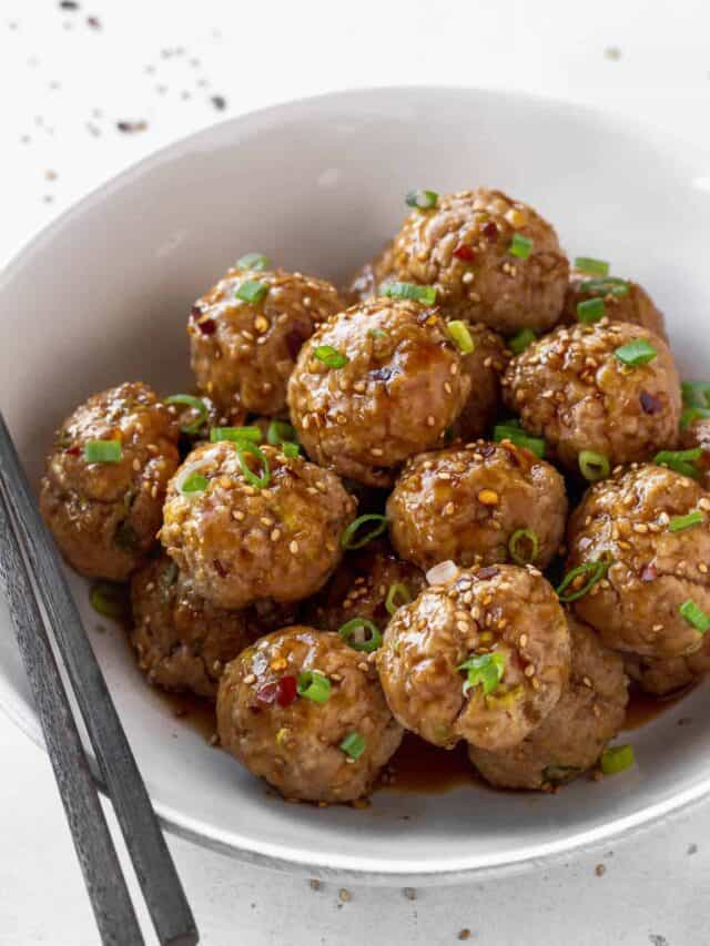 white serving bowl filled with asian pork meatballs with sweet soy dipping sauce garnished with scallions and red pepper flakes.