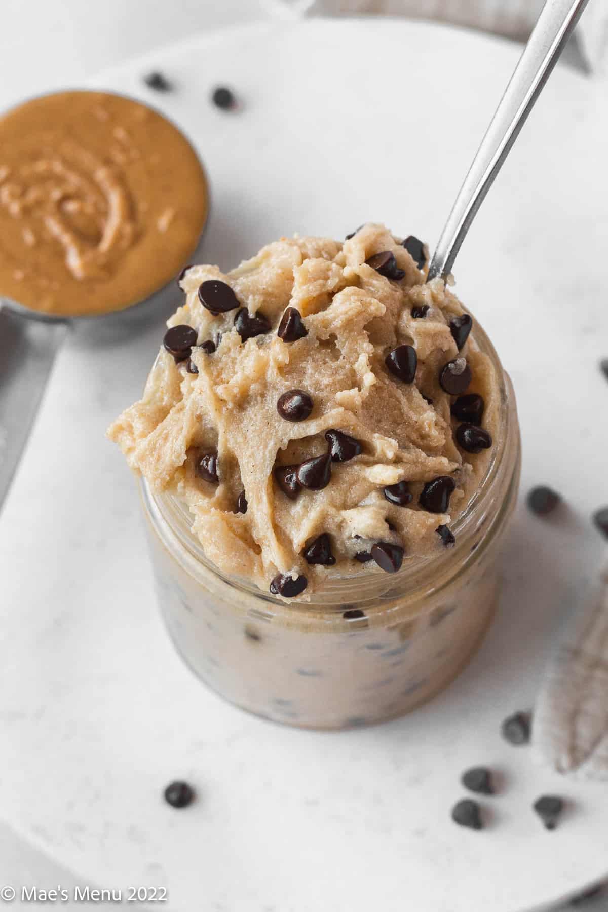 overhead shot of a glass jar of edible peanut butter chocolate chip cookie dough with a spoon sticking out and a ramekin of peanut butter off to the side.