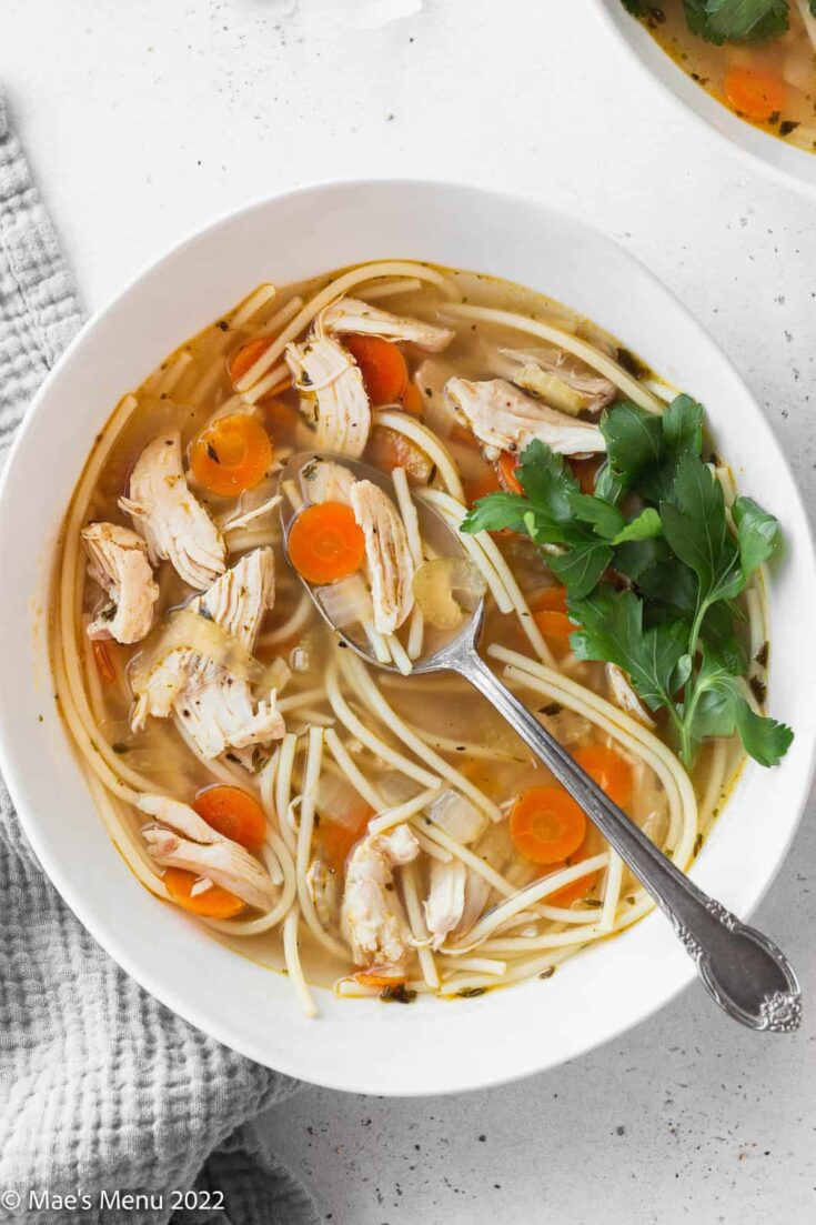 A large bowl of gluten-free chicken noodle soup with a silver spoon and a garnish of fresh parsley.
