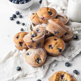 Dairy-free blueberry muffins piles on a dish cloth with blueberries, milk, and small flowers.