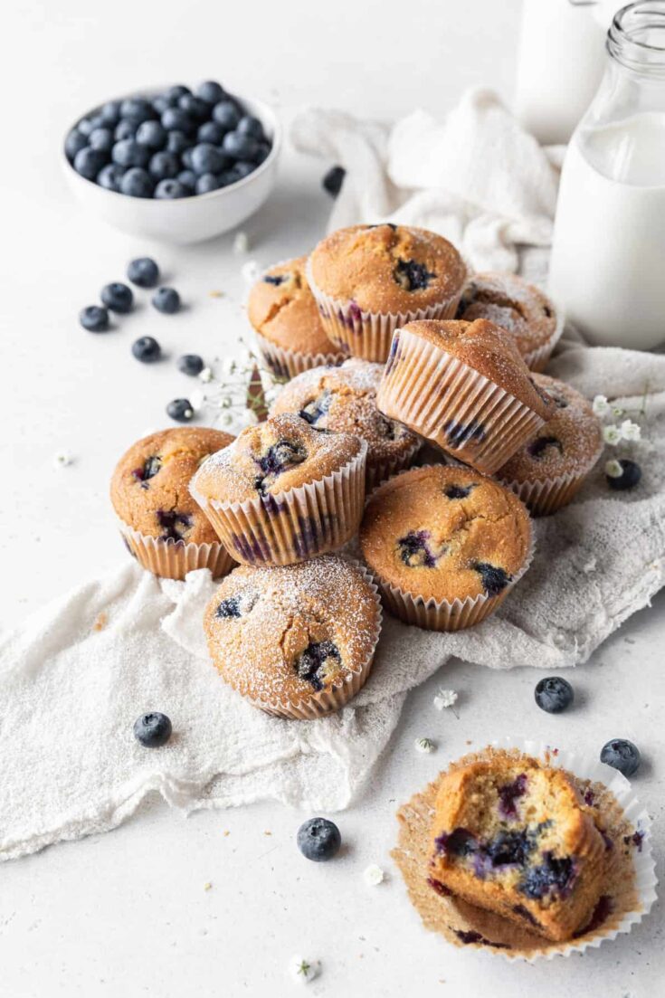 Dairy-free blueberry muffins piles on a dish cloth with blueberries, milk, and small flowers.