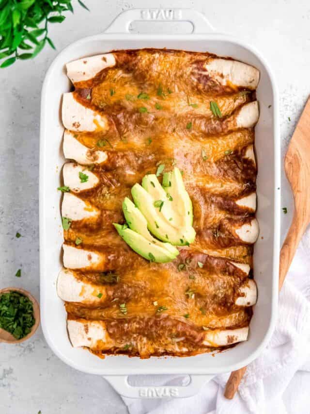 A white pan of ground turkey enchiladas with a serving spatula and cup of herbs on a white background.