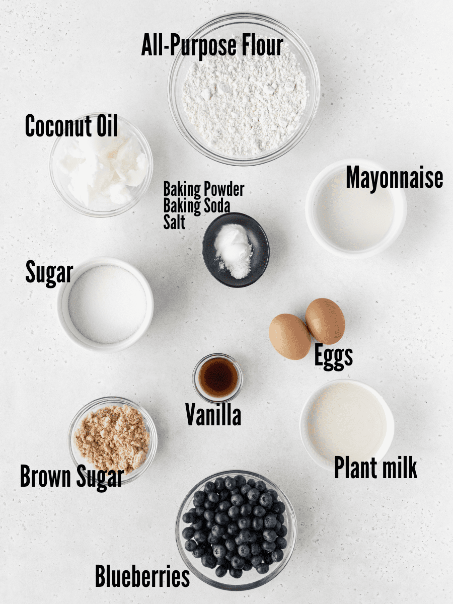 All of the ingredients for dairy-free blueberry muffins: blueberries, flour, sugar, brown sugar, plant milk, mayonnaise, vanilla, eggs, baking powder, baking soda, and salt.