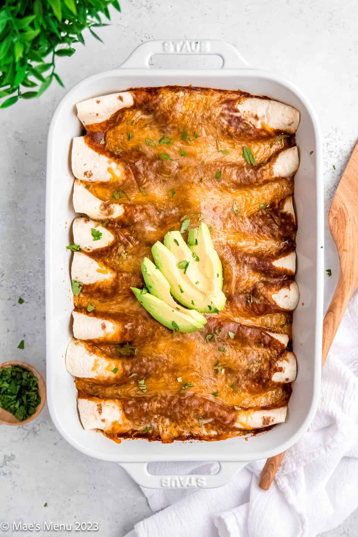 A white pan of ground turkey enchiladas with a serving spatula and cup of herbs on a white background.