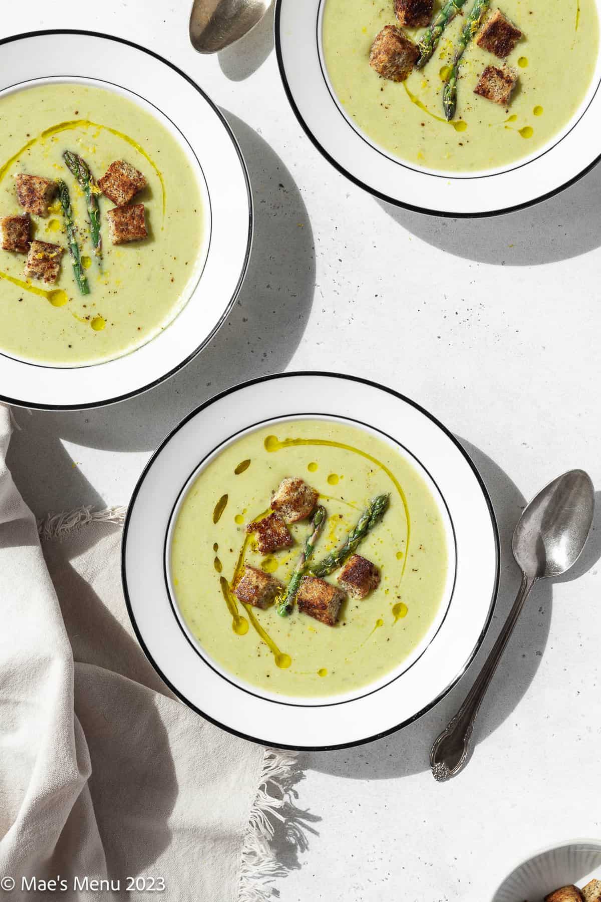 Three bowls of vegan asparagus soup on a white background with spoons and a towel.