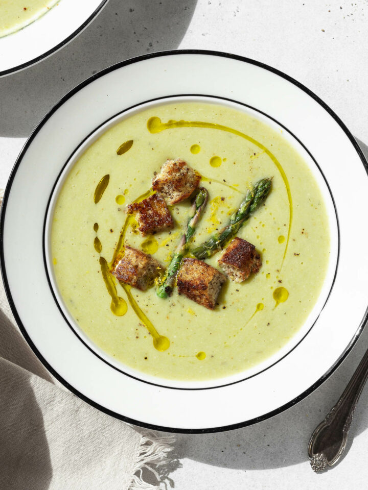 A large white bowl of asparagus soup without cream that is garnished with olive oil, croutons, asparagus, and black pepper.