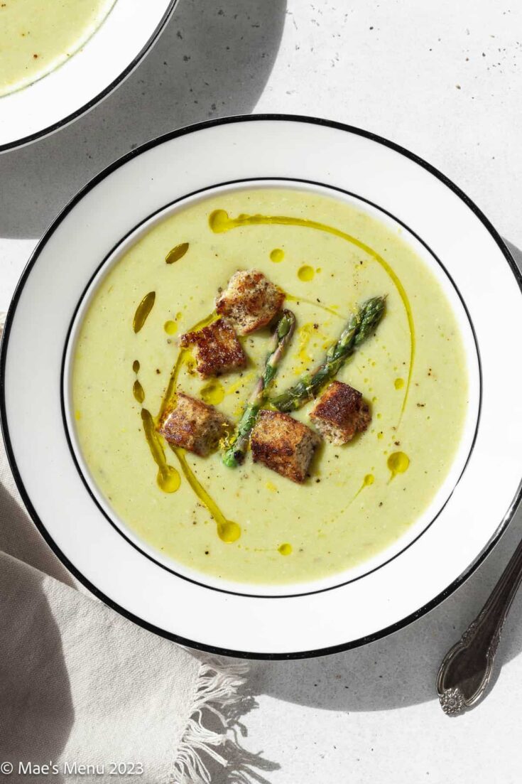 A large white bowl of asparagus soup without cream that is garnished with olive oil, croutons, asparagus, and black pepper.