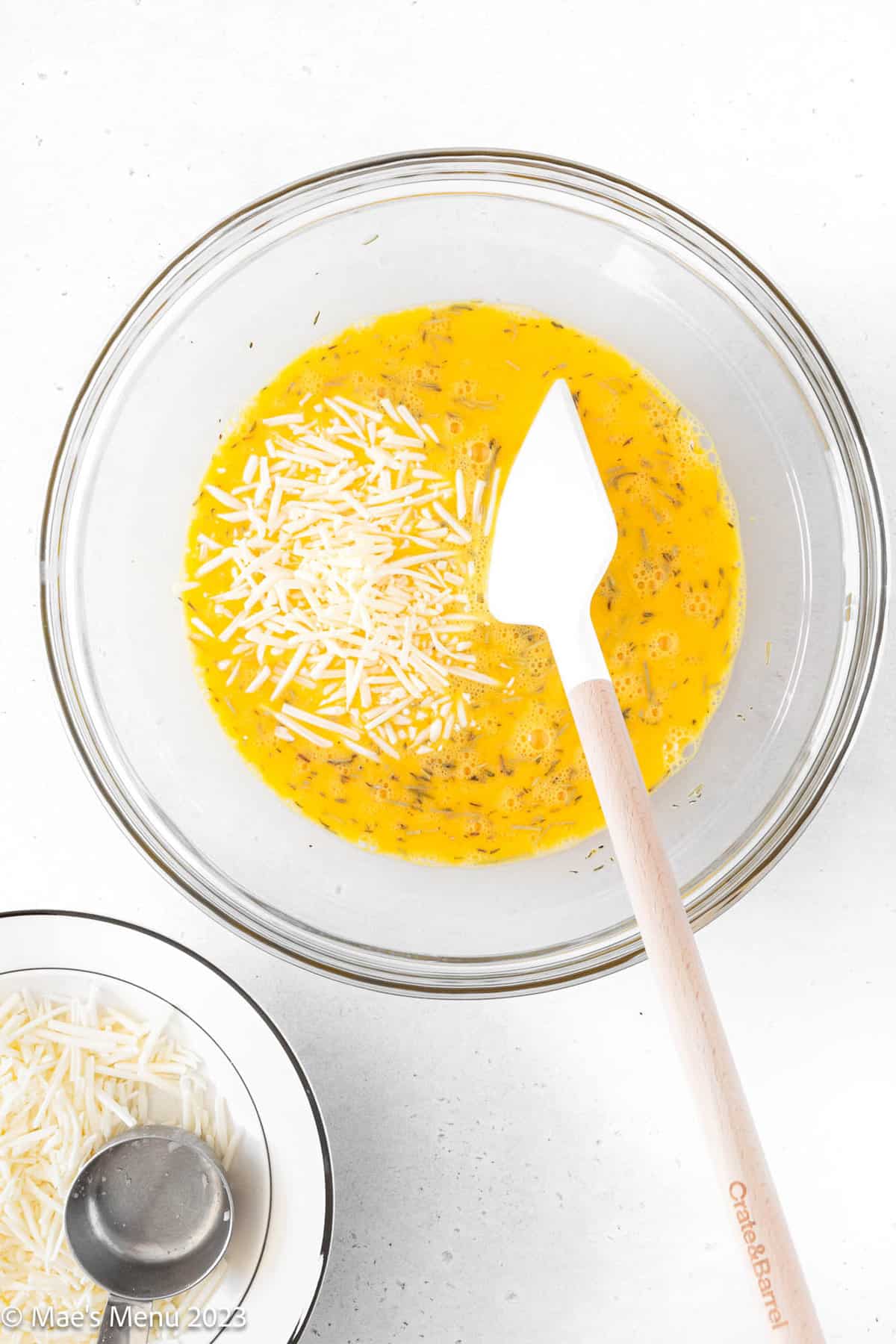 A glass mixing bowl of eggs and cheese with a spatula on a white background.