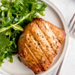 An overhead shot of a small dinner plate of grilled pork chops and arugula salad with a fork.