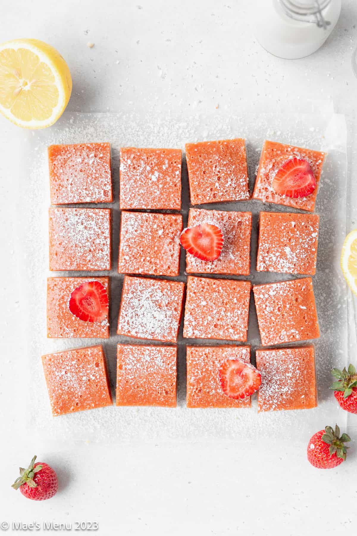 A cut up batch of strawberry lemonade bars on the counter with strawberries and lemons.