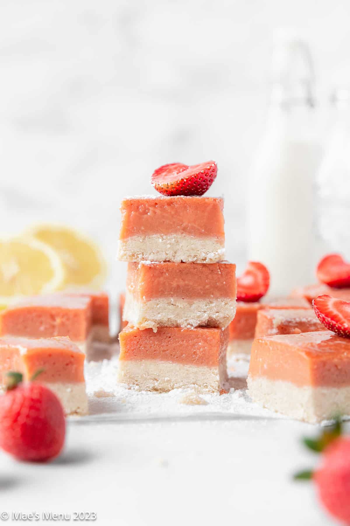 Stacks of strawberry lemonade bars on the counter with lemons, strawberries, and milk.