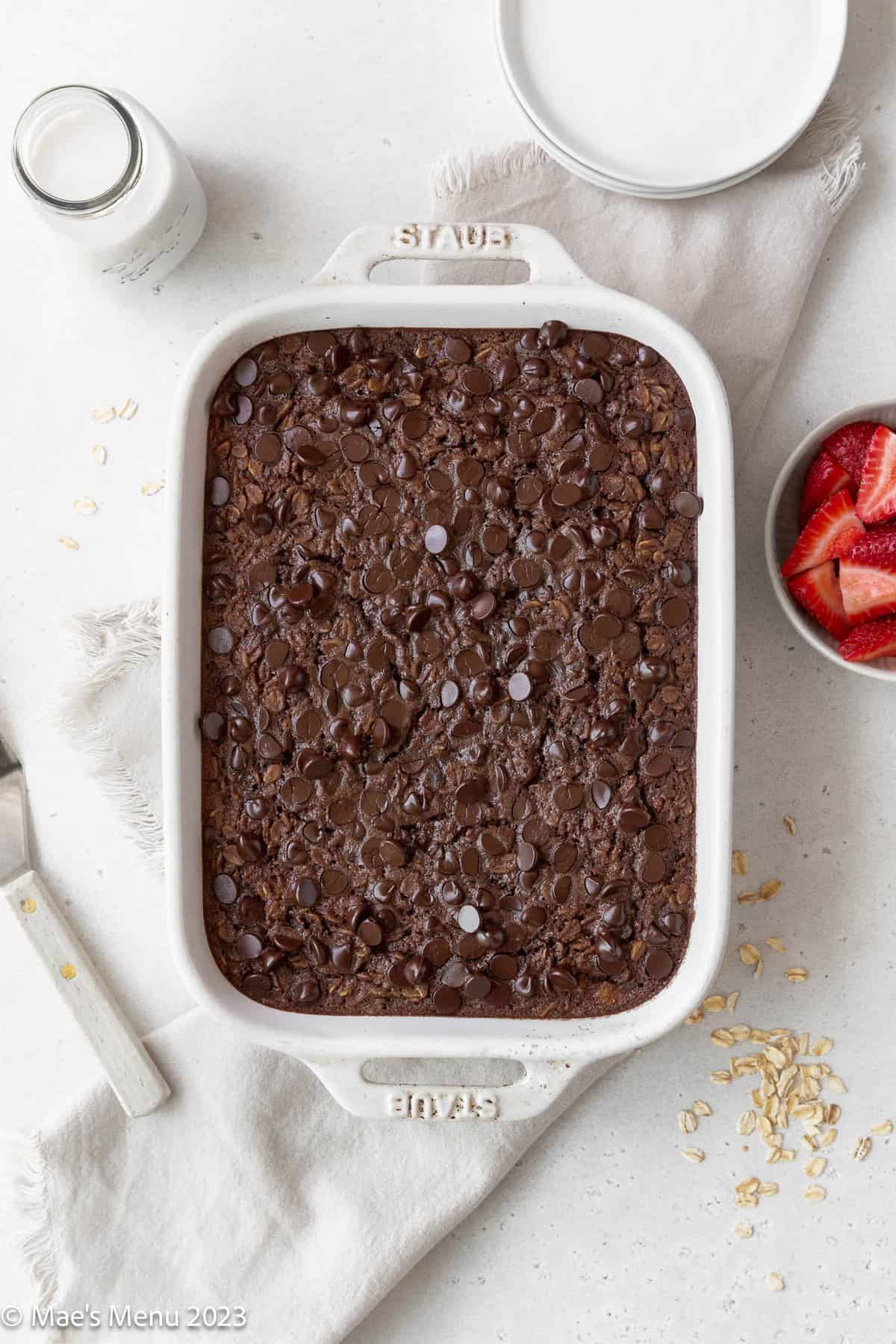 A large white pan of brownie baked oatmeal next to a bowl of strawberries, stack of white plates, oats, and jar of milk.