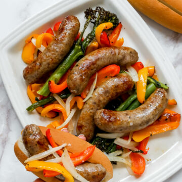 A hand holding an air fryer Italian sausage in a bun over the serving plate of the sausages and peppers.