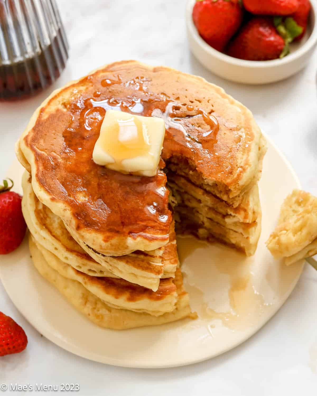 An overhead shot of a stakc of fluffy buttermilk pancakes with maple syrup on top and a wedge cut out of them.