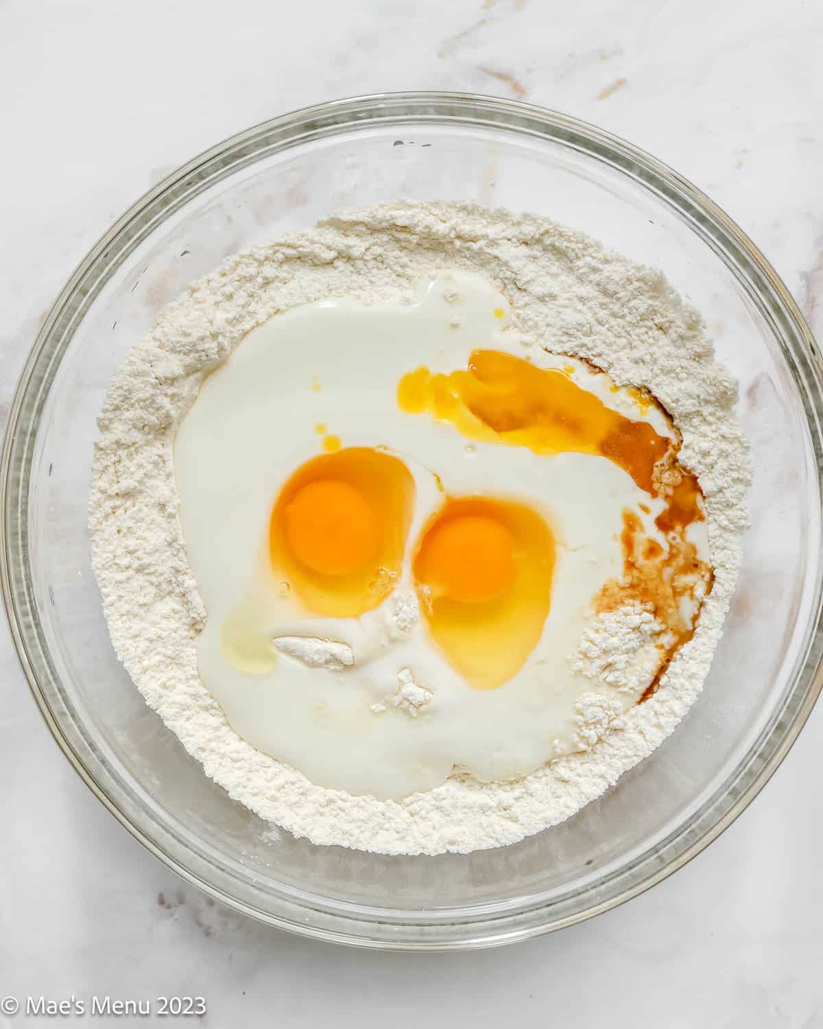 A glass mixing bowl with flour, milk, eggs, and vanilla.