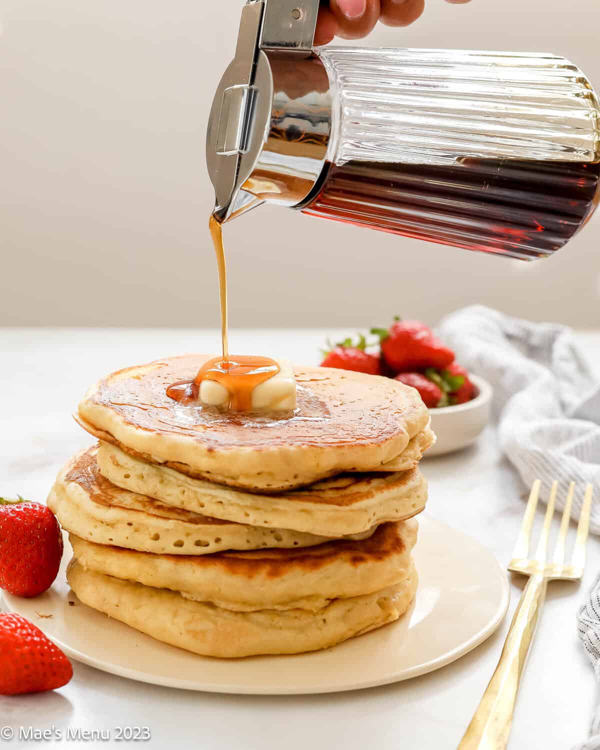 Drizzling maple syrup over a stack of fluffy buttermilk pancakes on a plate with strawberries.