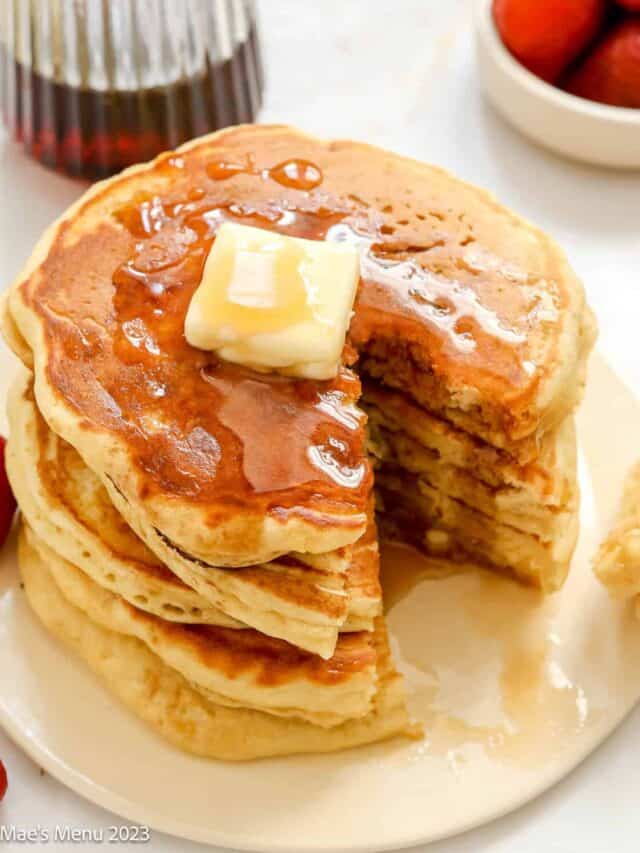 An overhead shot of a stack of fluffy buttermilk pancakes with a wedge cut out of them and maple syrup and butter on top.
