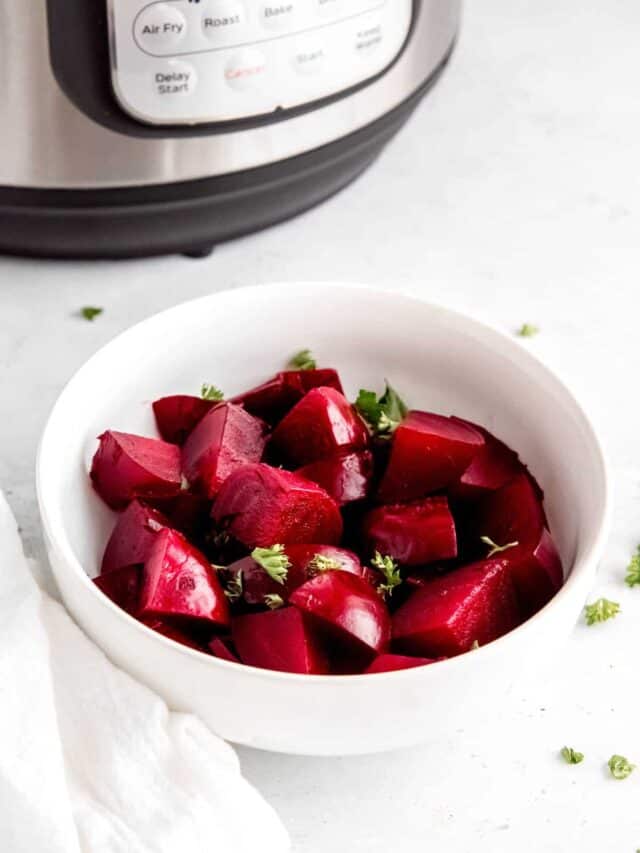 A white bowl of cooked beets with chopped curly parsley on the counter in front of the Instant Pot.
