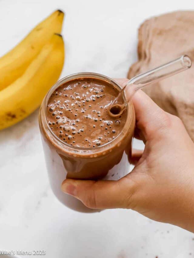 A hand holding up a glass of a mocha protein shake with a glass straw.