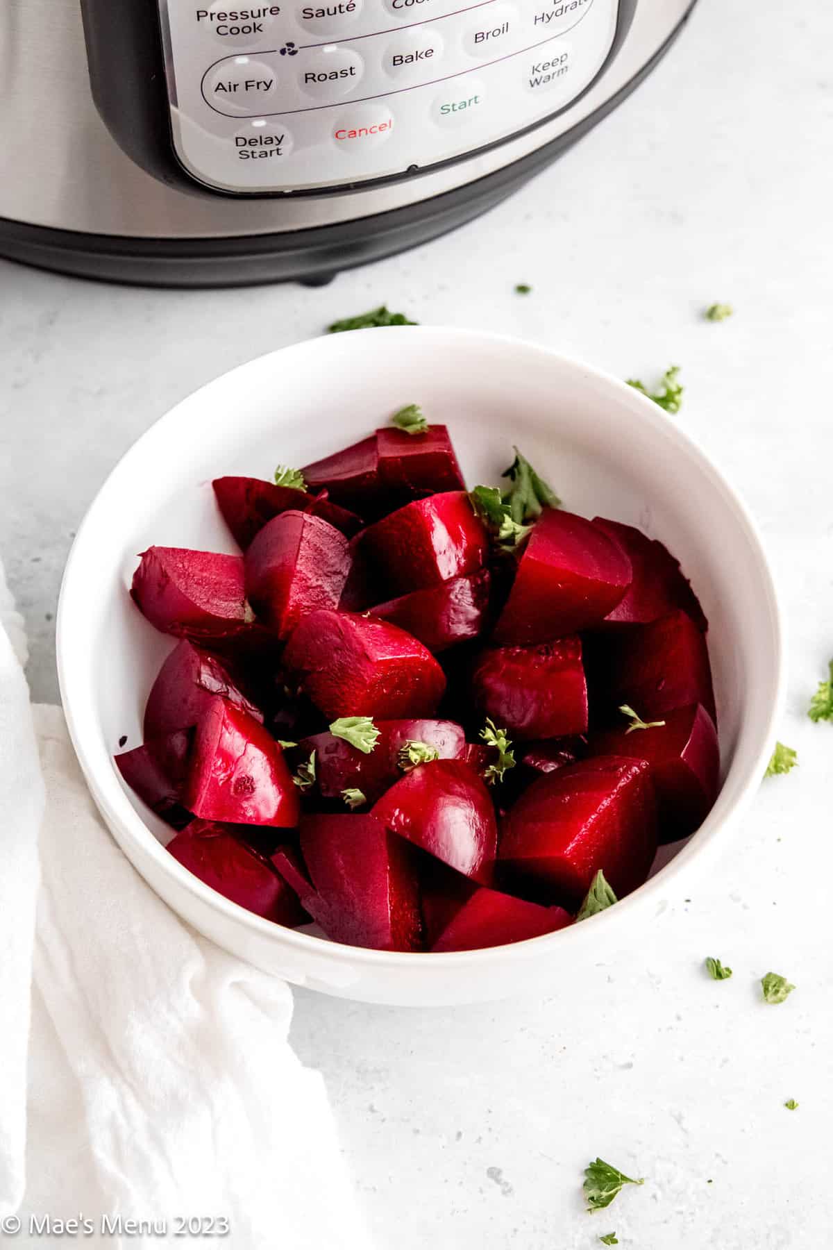 A white bowl of cooked cut up beets with curly parlsey on a white counter.
