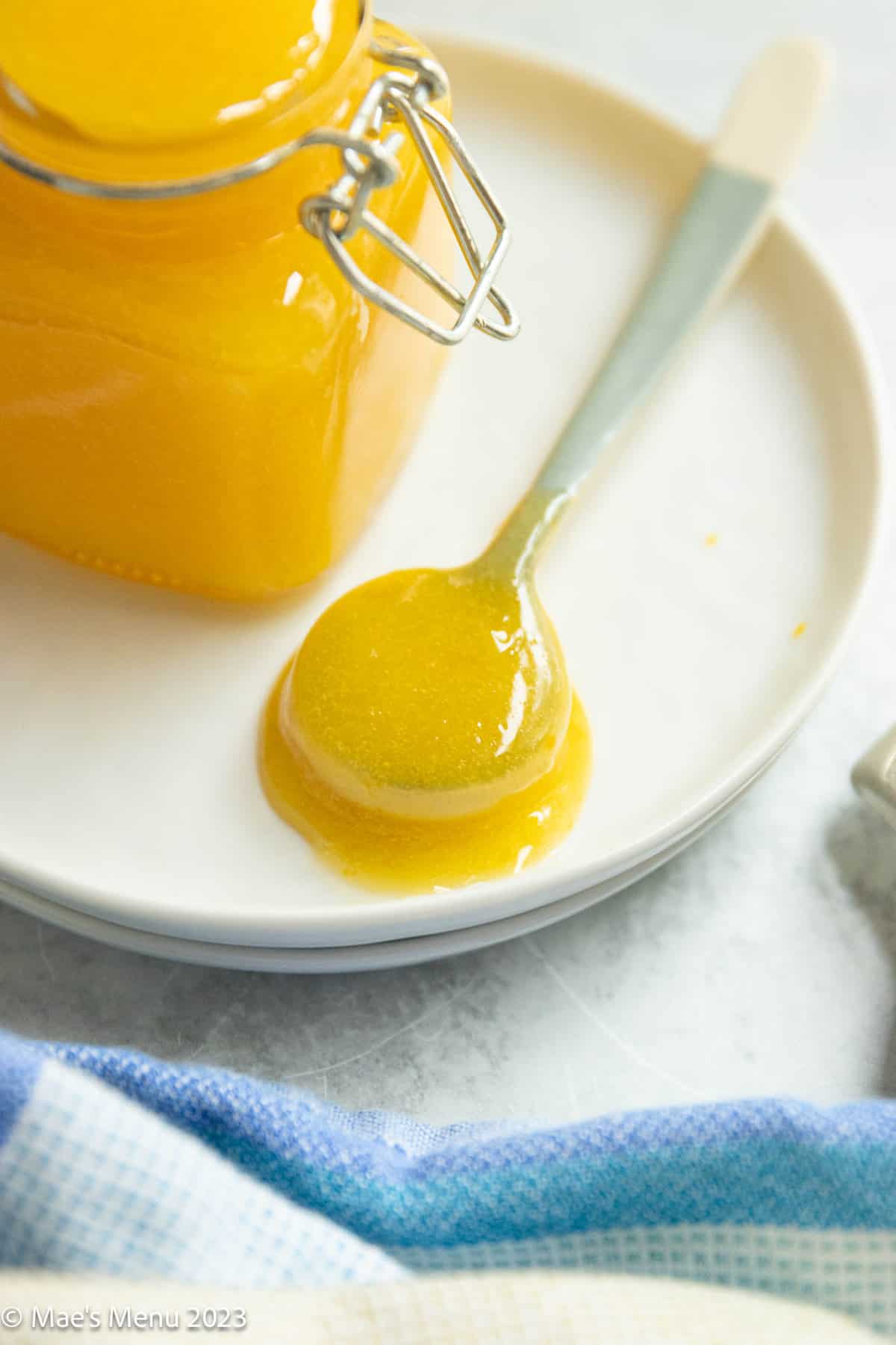 A small spoon of meyer lemon curd on a white plate next to a towel and a pot of the curd.